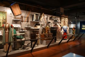 A brightly lit museum exhibit with vintage costumes, musical instruments, machines, banners, and artifacts on display behind barriers. *AI generated alt text