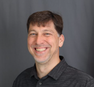 A smiling Italian American man with short brown hair and a beard stands in front of a plain gray background, wearing a dark collared shirt.