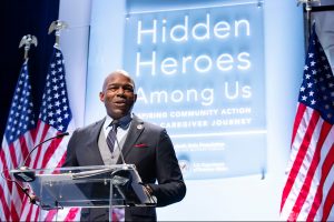 A man in a suit speaks at a clear podium with two American flags behind him and a backdrop with the text “Hidden Heroes Among Us: Inspiring Community Action & Caregiver Journey.”. *AI generated alt text