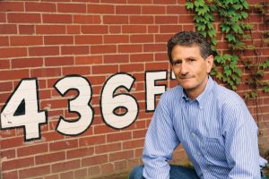 A man in a blue striped shirt sits before a red brick wall marked 436F, with green ivy covering the upper right area of the wall. *AI generated alt text