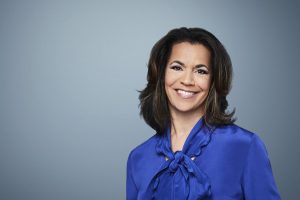 A woman with shoulder-length dark hair, in a bright blue blouse with a bow collar, smiles at the camera before a light blue-gray backdrop. *AI generated alt text