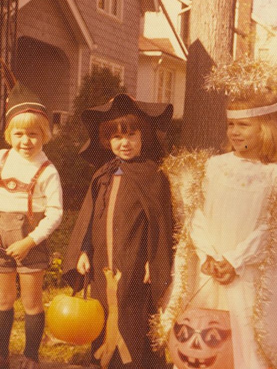 Four children in Halloween costumes, each with pumpkin candy buckets, stand outdoors; houses and trees visible behind them. *AI generated alt text