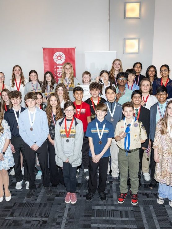 A diverse group of students with medals and badges on a stage at the National History Day awards; red banner and podium behind them. *AI generated alt text