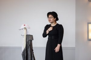 A person in glasses, black hat, and black clothes stands indoors with one hand on chest. A coat stand holds a nurse's hat and gray coat. *AI generated alt text