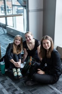 Three young women in black outfits sit on a carpeted floor by large windows, with bags and a green water bottle nearby. One has a green cast. *AI generated alt text