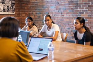 Three young women at a wooden table with laptops and water bottles, listening to another person, preparing for National History Day. *AI generated alt text