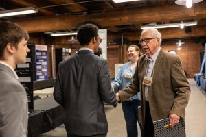 An older man in glasses shakes hands with a younger man in a suit at a National History Day event; people and posters are visible. *AI generated alt text