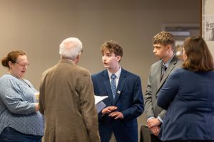 Five people, three in suits and two in casual dress, stand indoors discussing National History Day against a plain background. *AI generated alt text