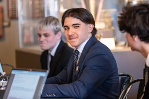 Three young men in suits sit at a table with laptops; one smiles at the camera while two others talk, in a conference room setting. *AI generated alt text