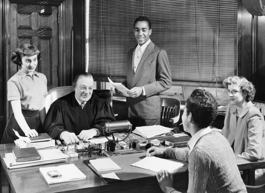 Black-and-white photo of a judge at his desk with four young adults holding act 48 papers in an office lined with books and papers. *AI generated alt text