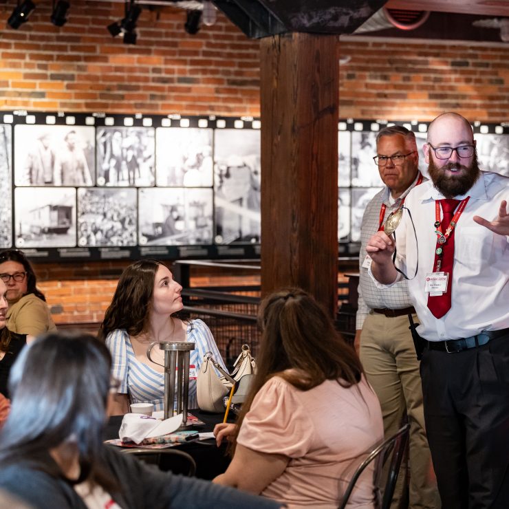 A man in a white shirt and red tie speaks to an attentive group in a brick-walled room with historic black-and-white photos. *AI generated alt text