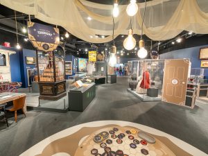 A well-lit US history museum exhibit with interactive displays, artifacts, display cases, vintage items, and a mannequin in period dress. *AI generated alt text