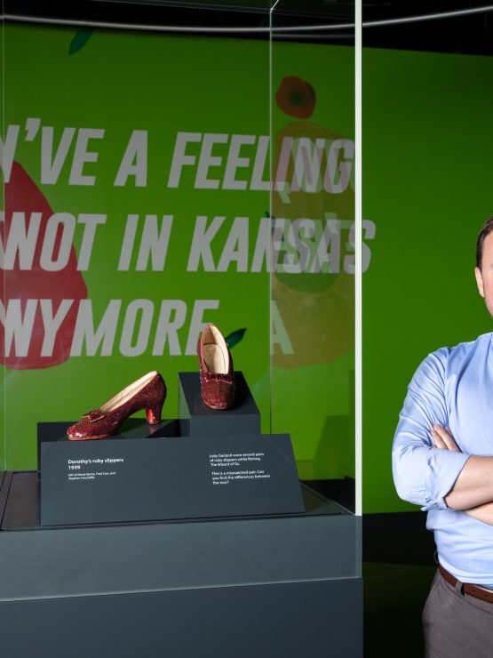 A man with arms crossed stands next to a glass case displaying ruby slippers at the Smithsonian; text on green wall behind him. *AI generated alt text