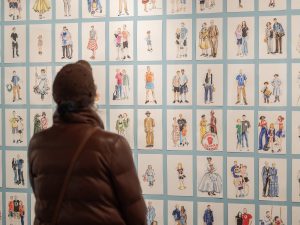 A person in a brown jacket and hat stands before a wall with many small, framed illustrations of people in historical attire and poses. *AI generated alt text