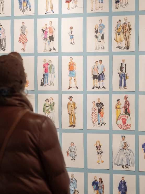 A person in a brown jacket and hat stands before a wall with many small, framed illustrations of people in historical attire and poses. *AI generated alt text