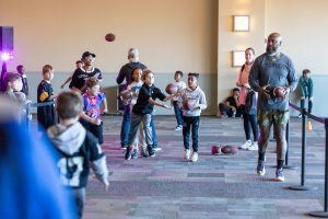 Children and adults take part in an indoor football throwing activity, aiming at a target, with a man in athletic wear in front. *AI generated alt text