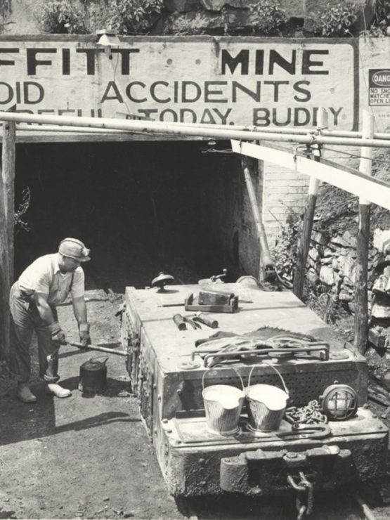 A miner stands by equipment outside Moffitt Mine; sign above reads “Moffitt Mine. Avoid Accidents. Be Careful Today, Buddy.”. *AI generated alt text