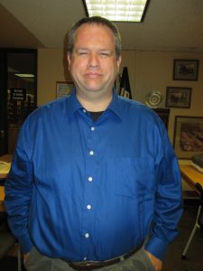 A man with short hair in a blue button-up shirt stands indoors, hands in pockets. Framed pictures and a trophy are in the background. *AI generated alt text