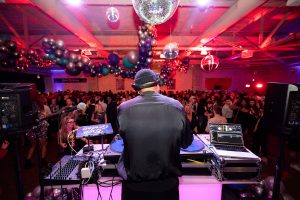 A DJ at an indoor party with colorful lighting, balloon decorations, and disco balls overhead. People are dancing on a crowded floor. *AI generated alt text