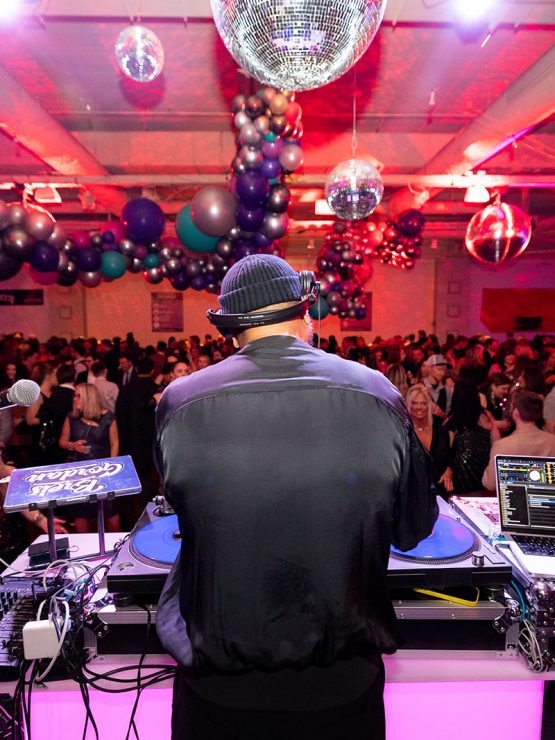 A DJ at an indoor party with colorful lighting, balloon decorations, and disco balls overhead. People are dancing on a crowded floor. *AI generated alt text