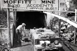 A miner stands by a rail cart at Moffitt Mine entrance, under a safety sign. Buckets, rocks, and mining tools are around him. *AI generated alt text
