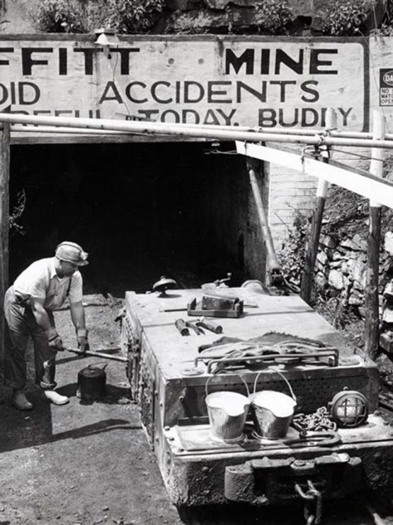 A miner stands by a rail cart at Moffitt Mine entrance, under a safety sign. Buckets, rocks, and mining tools are around him. *AI generated alt text