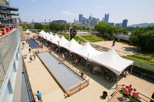 White event tents line a walkway next to a bocce court, with people gathered. City skyline and yellow bridge appear in the background. *AI generated alt text