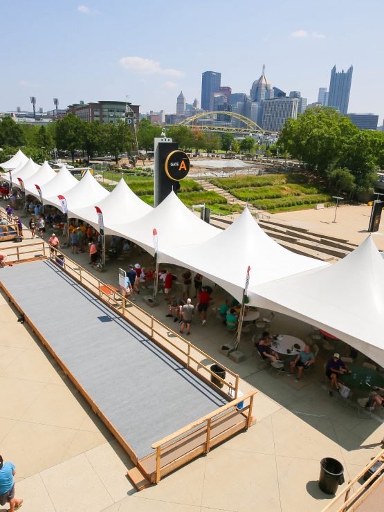 White event tents line a walkway next to a bocce court, with people gathered. City skyline and yellow bridge appear in the background. *AI generated alt text