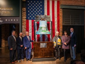 Six people pose indoors in front of a large Liberty Bell sculpture and American flag, with a brick wall and exhibit sign behind them. *AI generated alt text