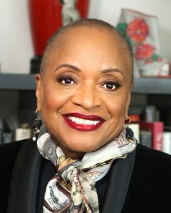 A woman with short hair, a patterned scarf, earrings, and red lipstick poses smiling in front of a blurred bookshelf. *AI generated alt text