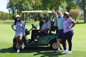 Five individuals on a golf course; two seated in a golf cart, three standing nearby, all in golf attire. Trees and grass behind. *AI generated alt text