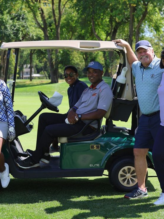 Five individuals on a golf course; two seated in a golf cart, three standing nearby, all in golf attire. Trees and grass behind. *AI generated alt text