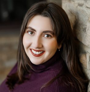 A woman with long brown hair in a plum turtleneck sweater leans against a stone wall, smiling. The background is softly blurred. *AI generated alt text