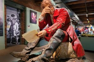 Life-sized statue of a man in a red colonial coat covering his face, holding a paper marked Act 48, among museum exhibits. *AI generated alt text