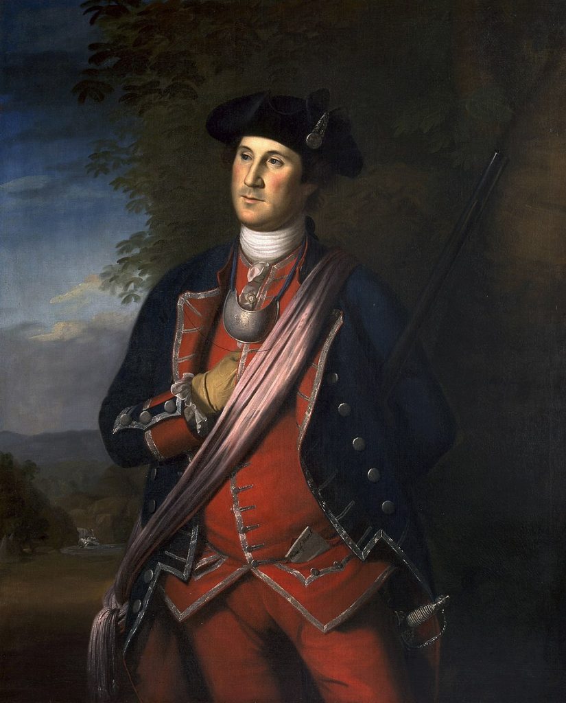 A man in 18th-century military uniform stands outdoors with a rifle, wearing a dark blue coat with red accents, trees and mountains behind. *AI generated alt text