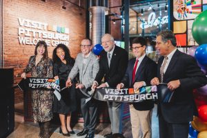 Six people at a ribbon-cutting in a museum, holding large scissors, with balloons and a VISIT PITTSBURGH WELCOME CENTER sign behind them. *AI generated alt text