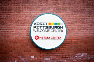 A round sign on a red brick wall reads Visit Pittsburgh Welcome Center and displays the history museum logo with Smithsonian below. *AI generated alt text
