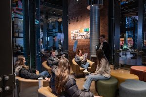 Six visitors sit and converse on modern chairs in a lobby area by a Welcome Center sign, with city views through large windows. *AI generated alt text