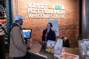 A man talks with a staff member at a museum information desk, with brochures and a computer in front of a brick wall with signage. *AI generated alt text