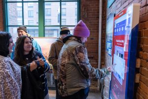 Visitors stand indoors by a brick wall as one person in a pink beanie uses an interactive kiosk; daylight streams through a large window. *AI generated alt text
