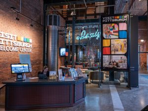 A person sits at the front desk of the history museum’s welcome center, with brochures, brick walls, and museum signage visible around them. *AI generated alt text