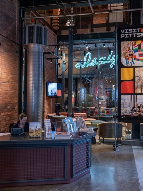 A person sits at the front desk of the history museum’s welcome center, with brochures, brick walls, and museum signage visible around them. *AI generated alt text
