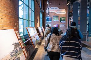 Visitors walk through a history museum exhibit with large photos, a sign on the brick wall, and big windows letting in sunlight. *AI generated alt text