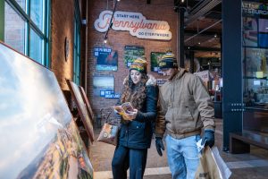 A woman and man in winter jackets and hats stand indoors, viewing a brochure near a large sign in a museum visitor center. *AI generated alt text