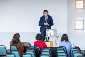 A person in a suit presents at the front of a room to three seated people with laptops. A small table with a device is nearby. *AI generated alt text