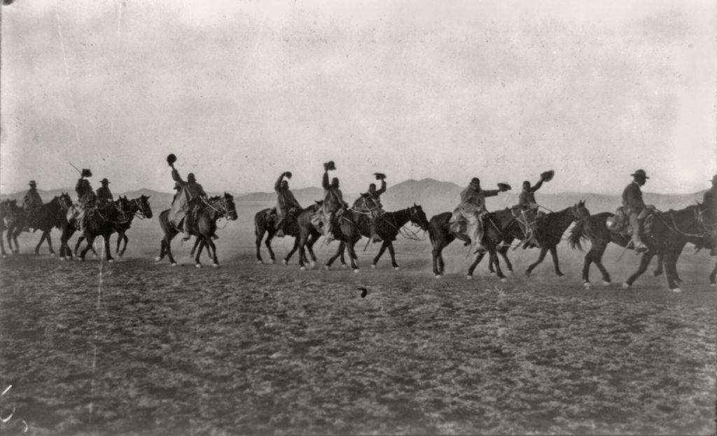 Black and white photo of horseback riders crossing a barren landscape, raising hats; faint mountains in the background. *AI generated alt text
