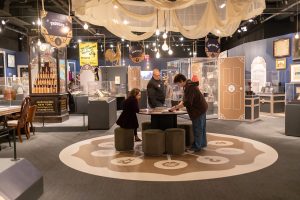 Three visitors interact with a circular table display surrounded by artifacts, signs, and draped ceiling fabric at a history museum exhibit. *AI generated alt text