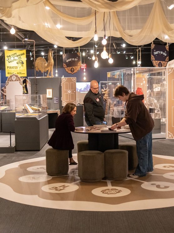 Three visitors interact with a circular table display surrounded by artifacts, signs, and draped ceiling fabric at a history museum exhibit. *AI generated alt text