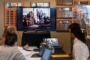 Four people at a table with laptops and papers view a historical painting on a large screen in a room with history award plaques. *AI generated alt text
