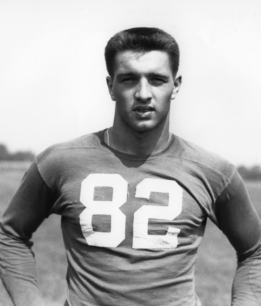 Black and white photograph of a young man in a long-sleeve football jersey numbered 82, standing outside on a field, expression neutral. *AI generated alt text